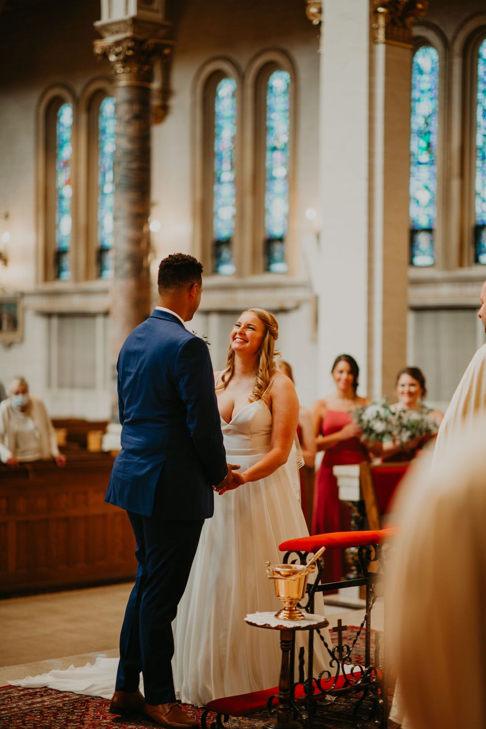 Bride and groom hold hands and smile at each other during church wedding with bridesmaids in background.