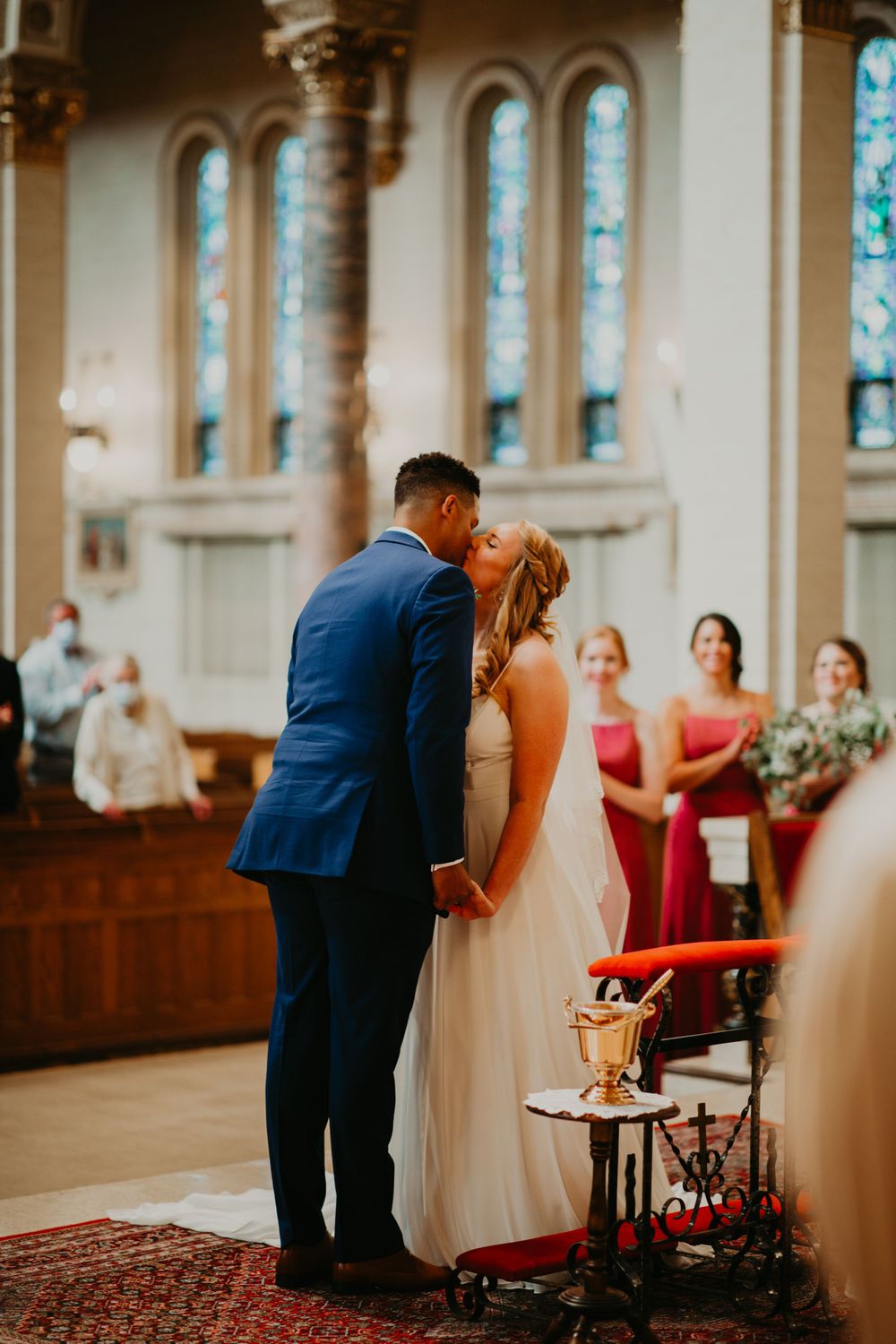 Newlyweds share first kiss at church altar as bridesmaids and guests look on during wedding ceremony.