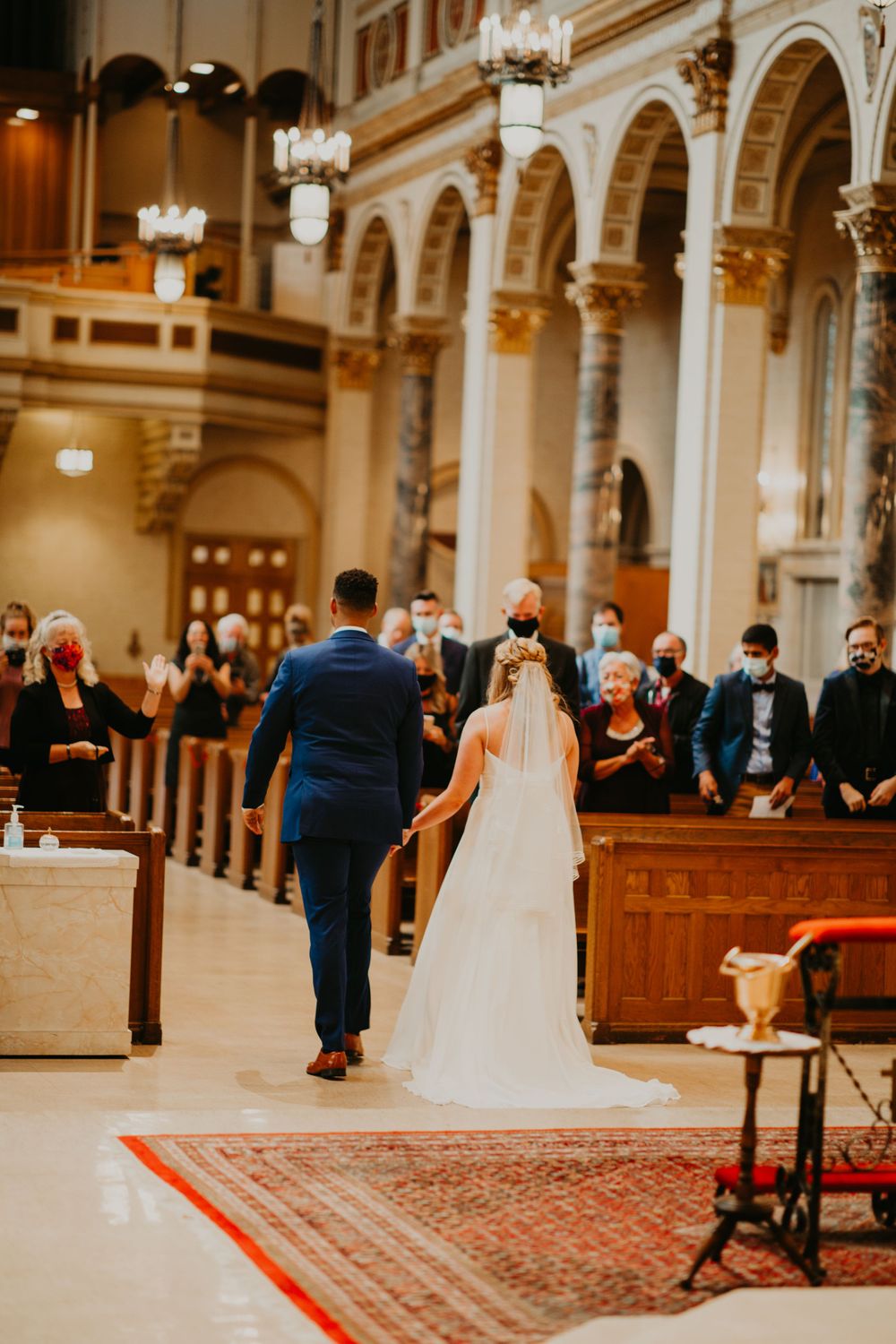 Newlyweds walk hand-in-hand down church aisle past masked guests after wedding ceremony.