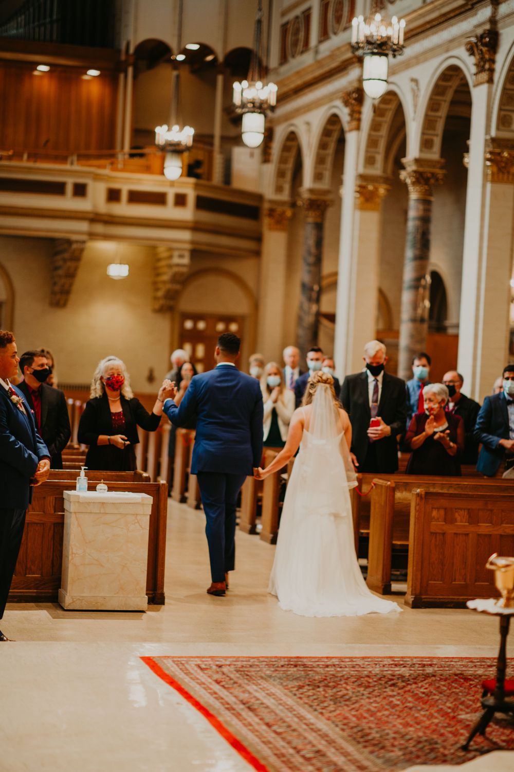 Bride and groom walk up church aisle holding hands as masked wedding guests stand in pews.