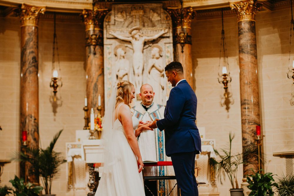 Bride and groom exchange rings at ornate church altar with smiling officiant during wedding ceremony.