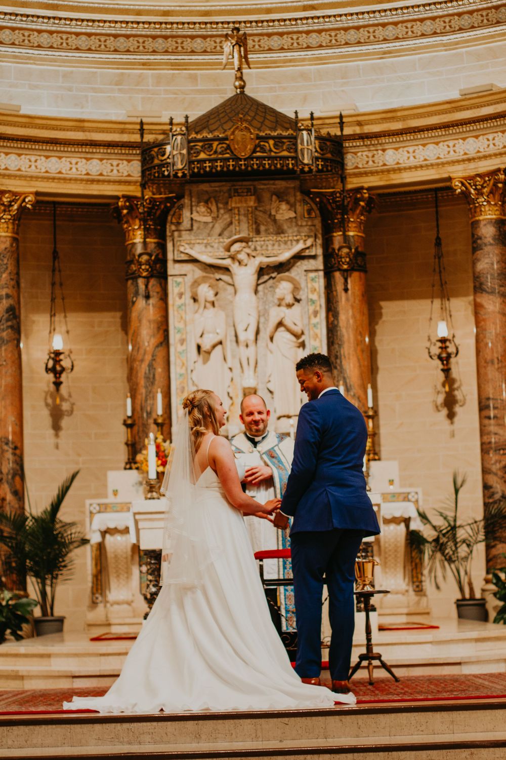 Couple holds hands before officiant at ornate church altar during wedding ceremony.