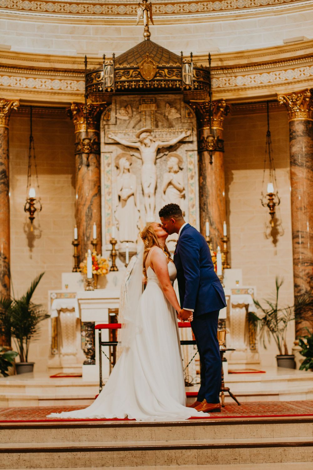 Newlyweds share first kiss at elegant church altar with decorative crucifix behind them.
