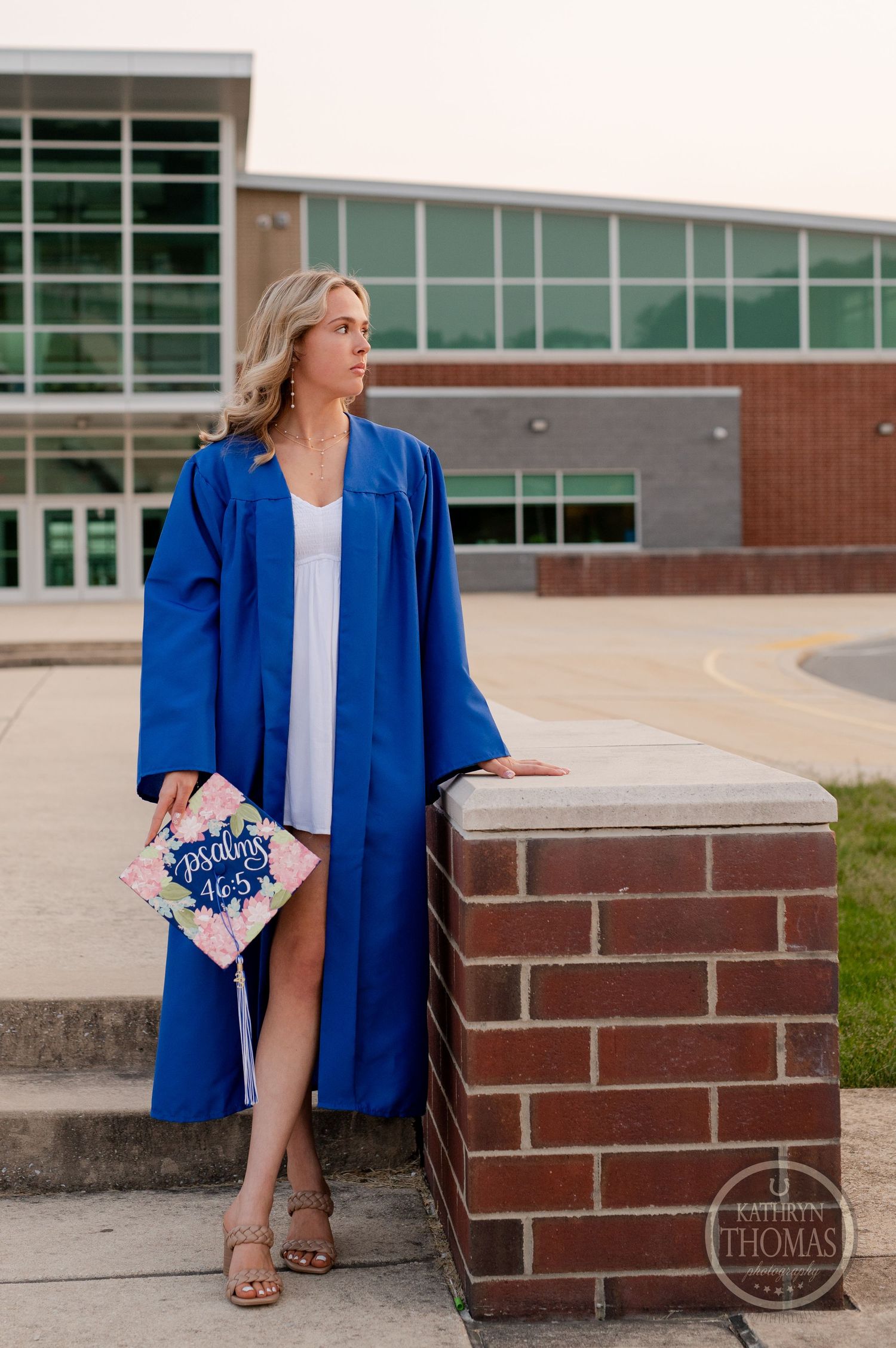 Graduate in blue gown holds decorated cap while posing against brick wall.