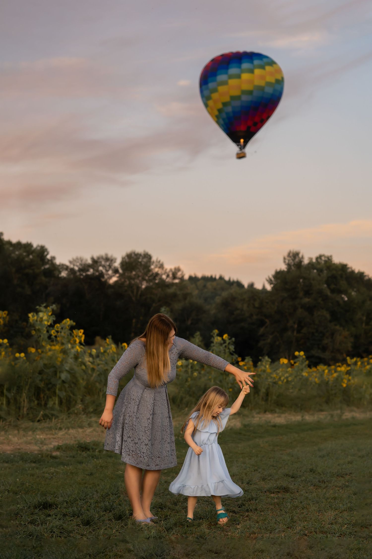 Hot air balloon floating over a sunflower field at sunset while people play in the foreground.