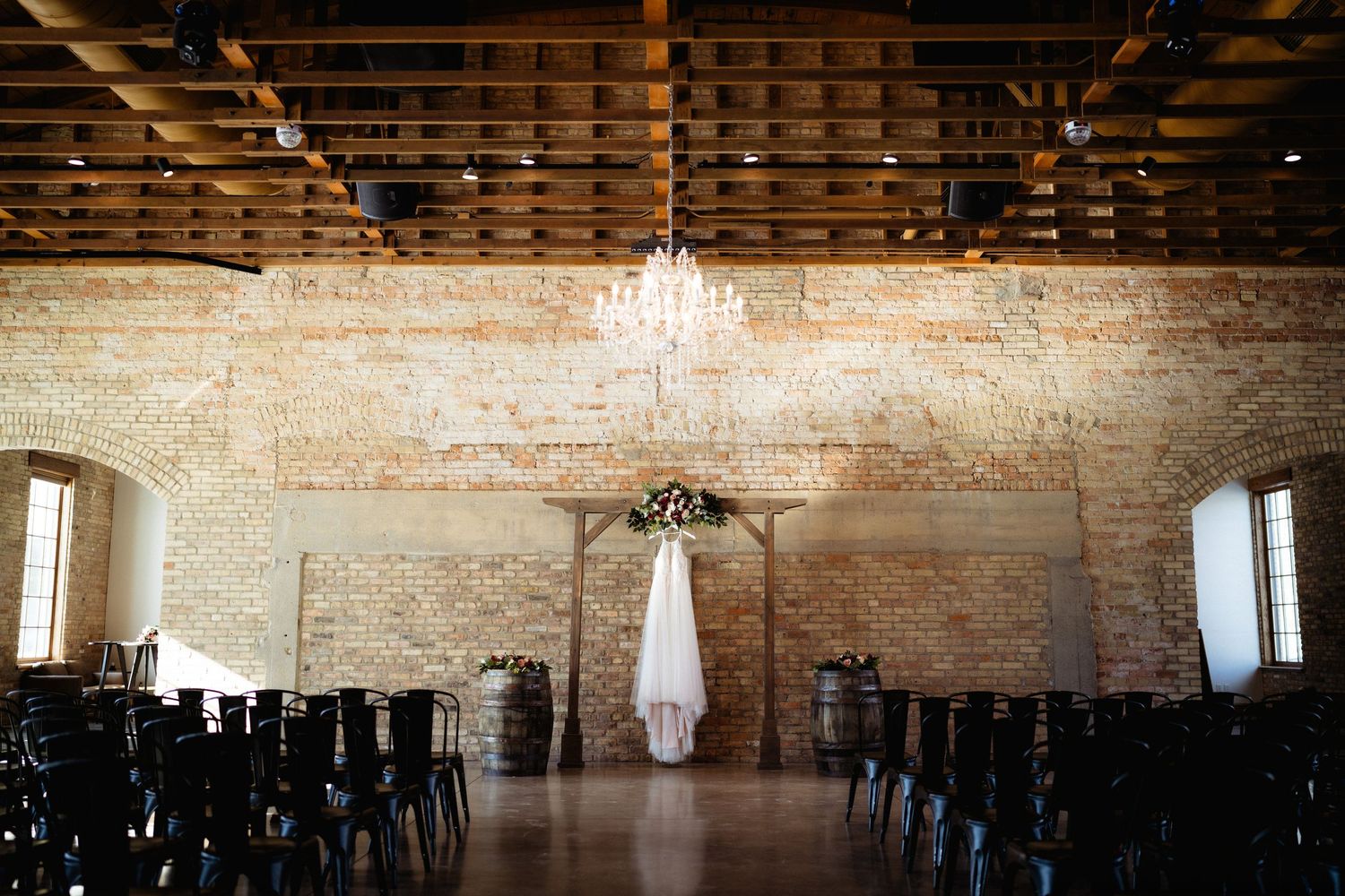Wedding dress hangs from a chandelier in a rustic brick venue with wooden chairs.