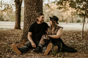 A couple sits beneath autumn trees on fallen leaves in a park setting.