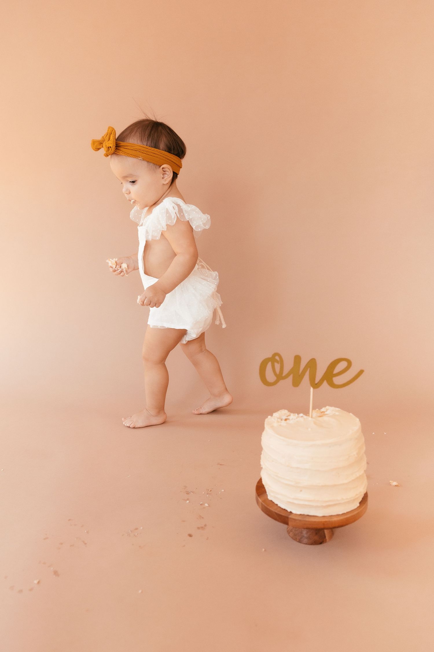 Baby in white outfit walking past birthday cake with gold 'one' topper.
