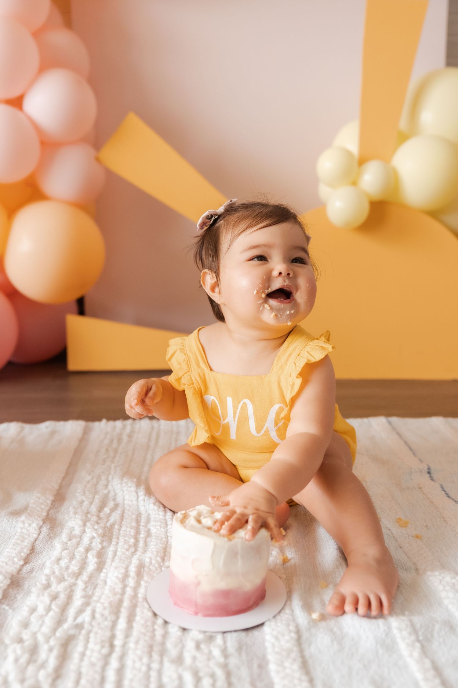 Baby in yellow outfit enjoying smash cake with balloon decorations.