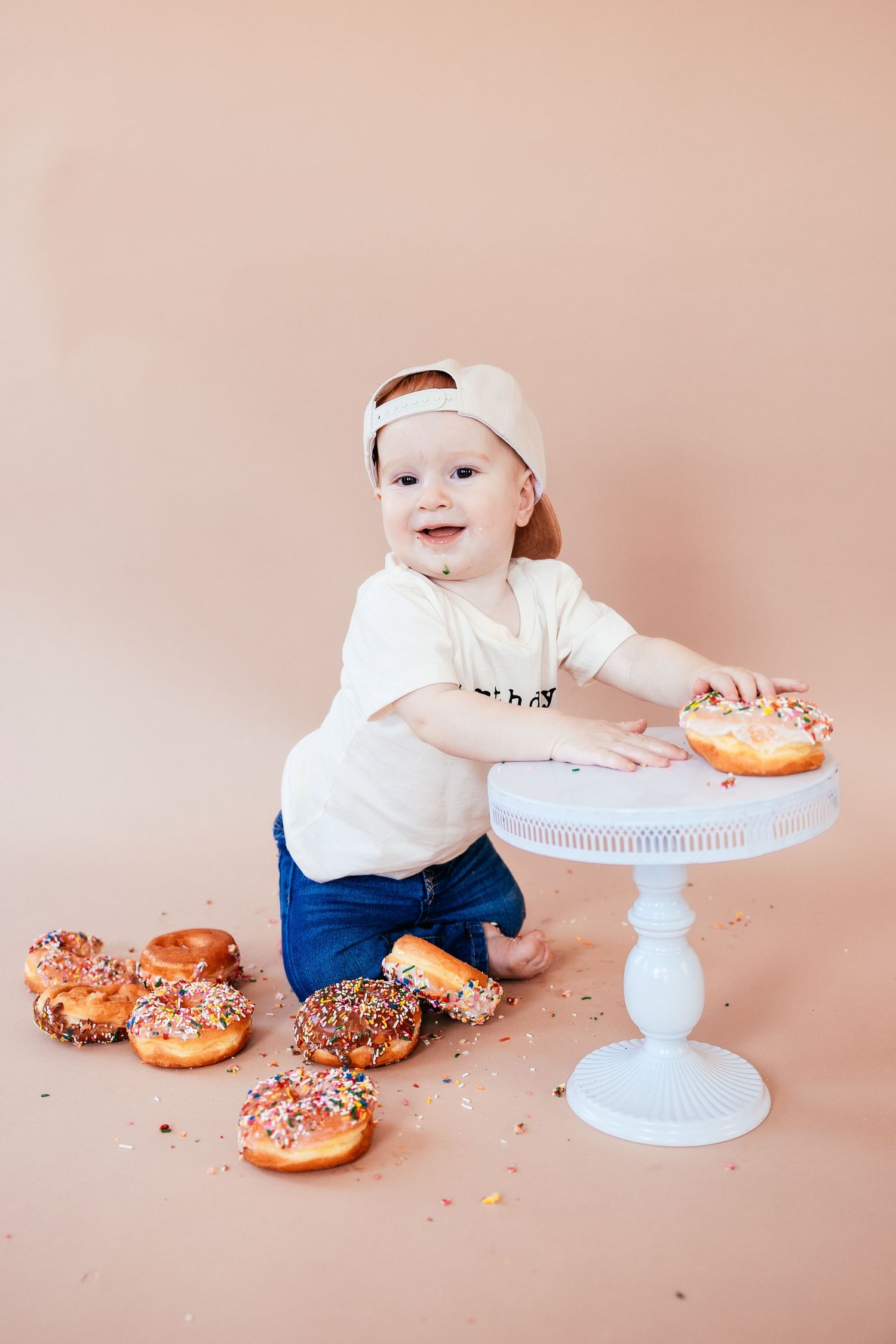 Toddler in white shirt and headband plays with colorful donuts on white cake stand.