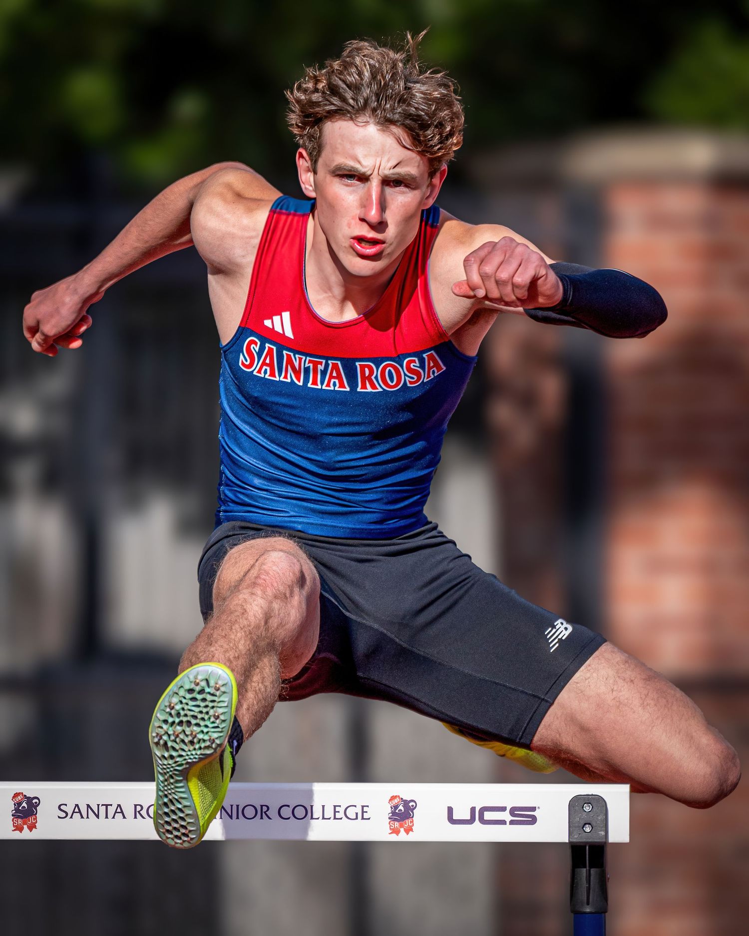 Track athlete leaping over hurdle in red and blue Santa Rosa uniform during competition.