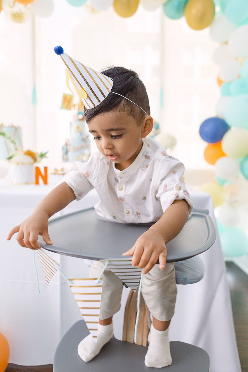 a one year old boy sits on a high chain in front of his birthday party decor in Hoboken photo by Paula Ortiz NY Family