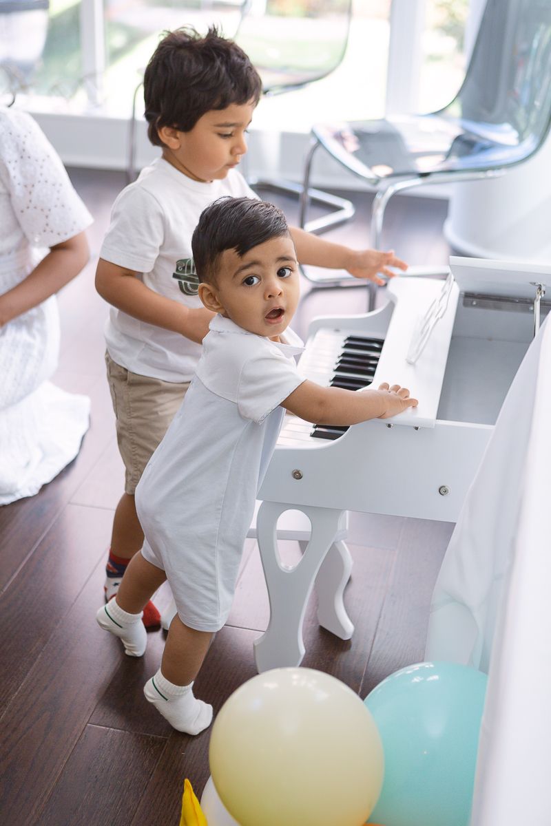 a one year old wearing light blue stands next to a small white piano with a friend on a candid moment by Paula Ortiz
