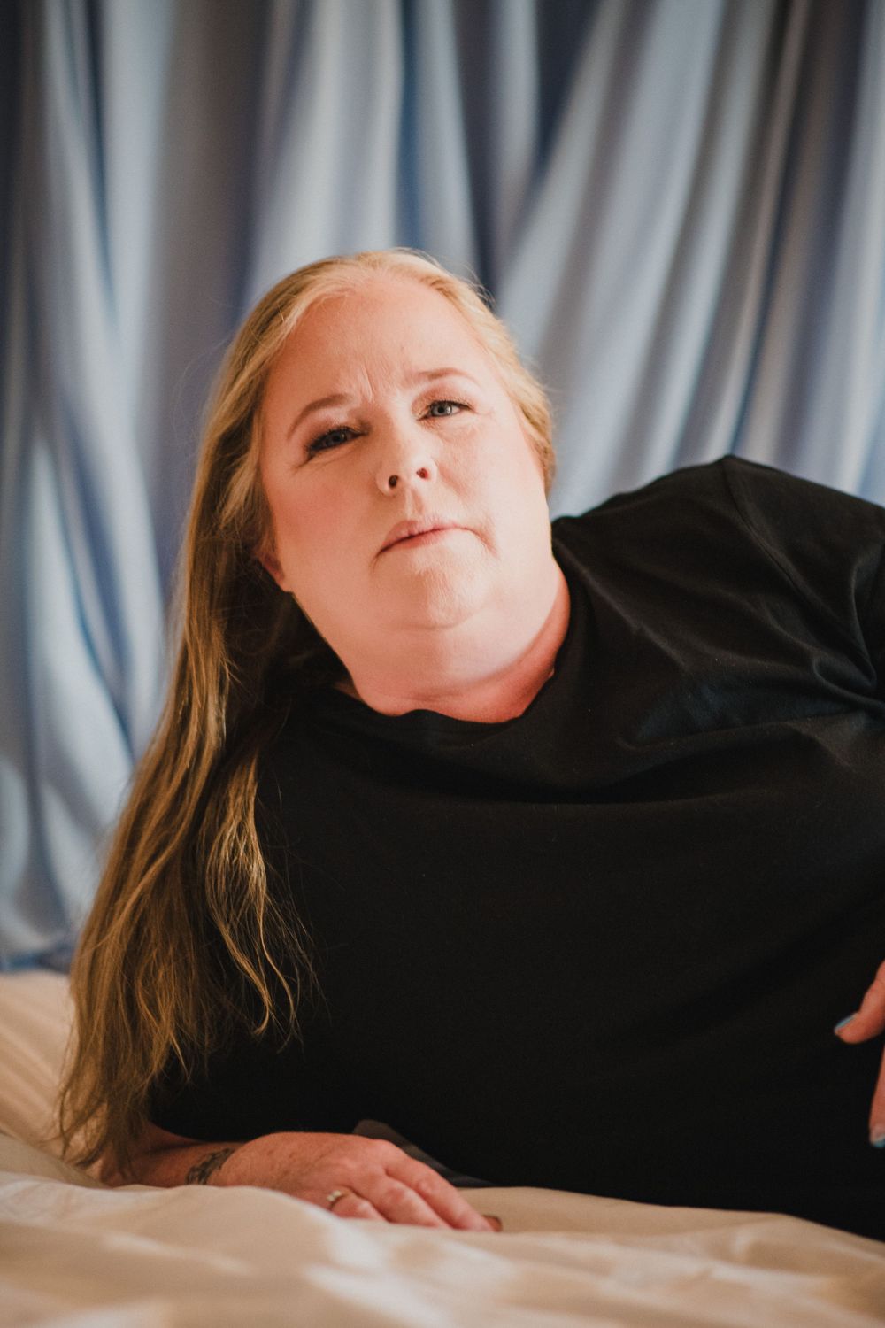 A person in a black top rests on light colored bedding against a draped backdrop.