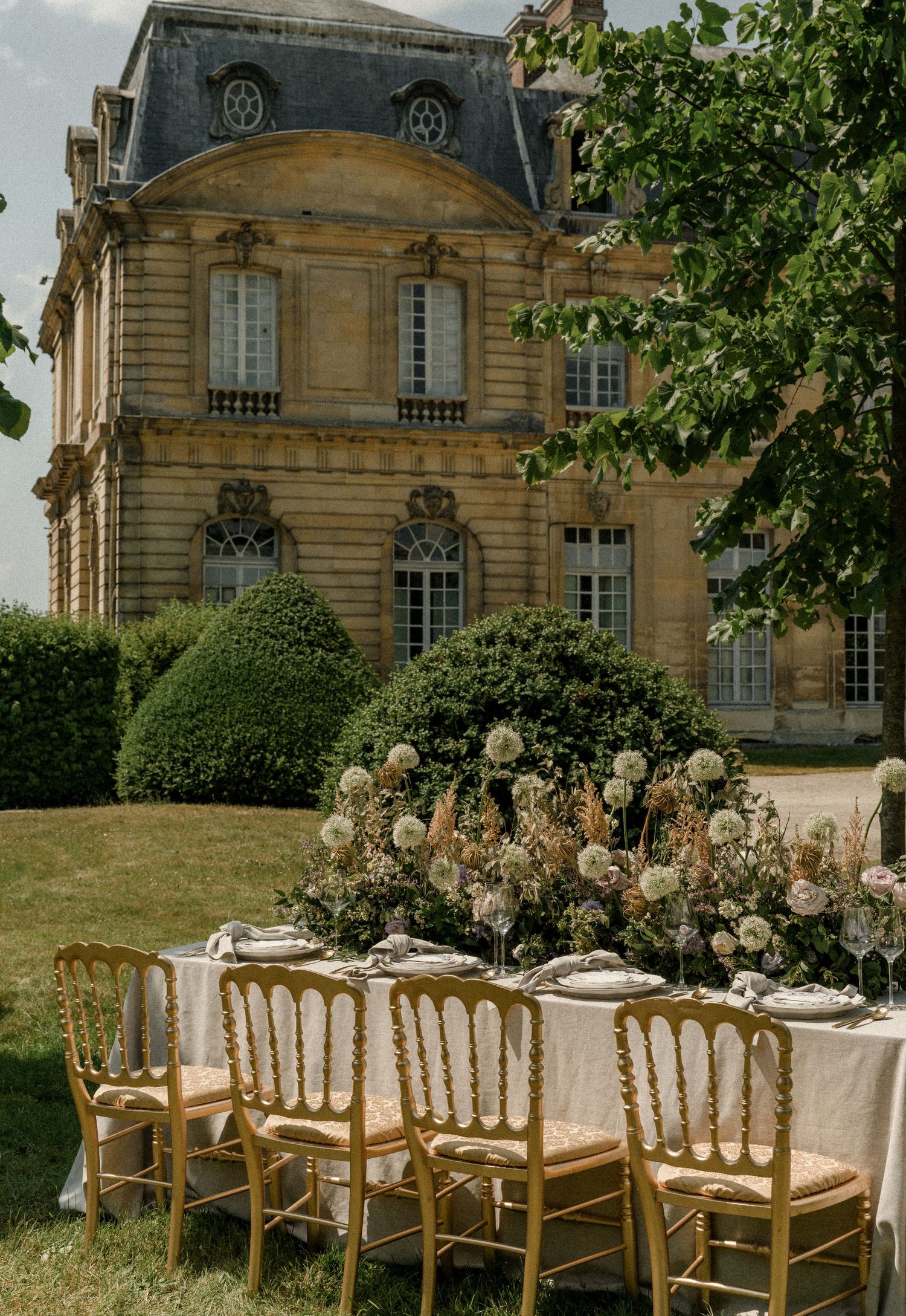 An elegant outdoor wedding table setting at a French chateau with gold chairs and floral centerpieces.