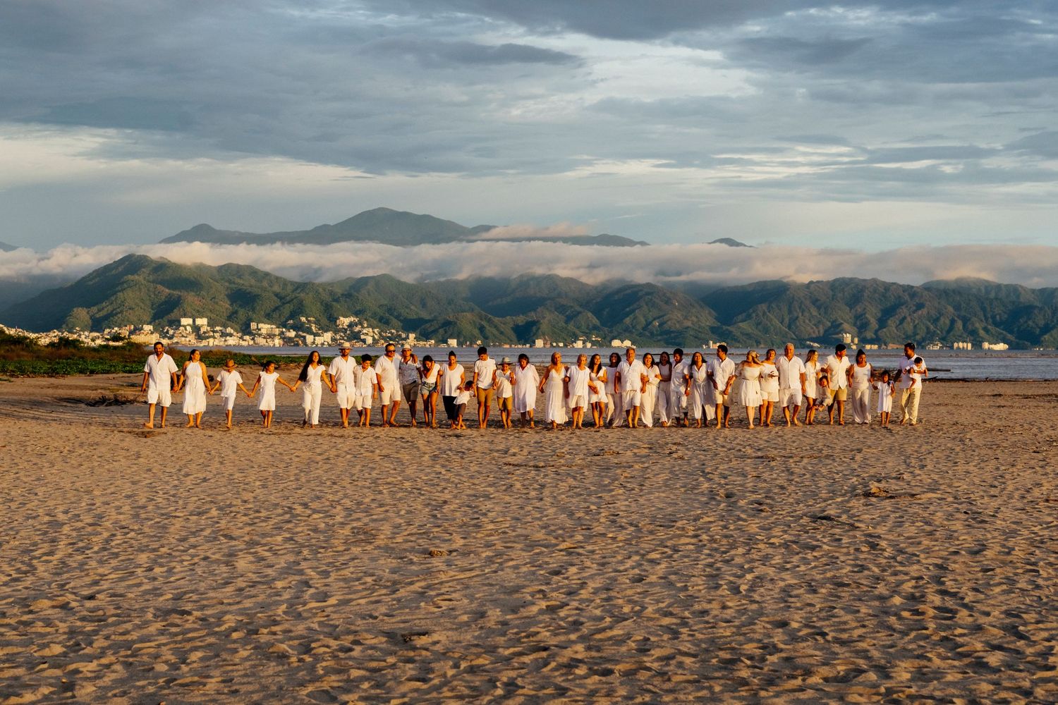 Large group photo of wedding guests gathered on a sandy beach with mountains in the distance.