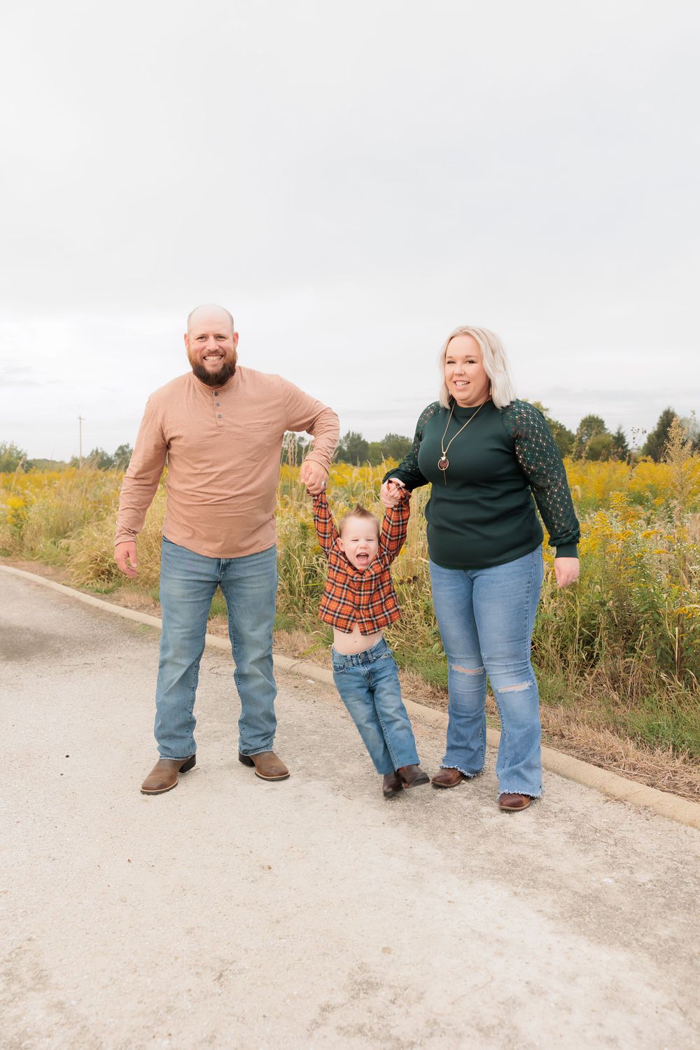 Family of three holding hands and jumping together on a path surrounded by autumn flowers.