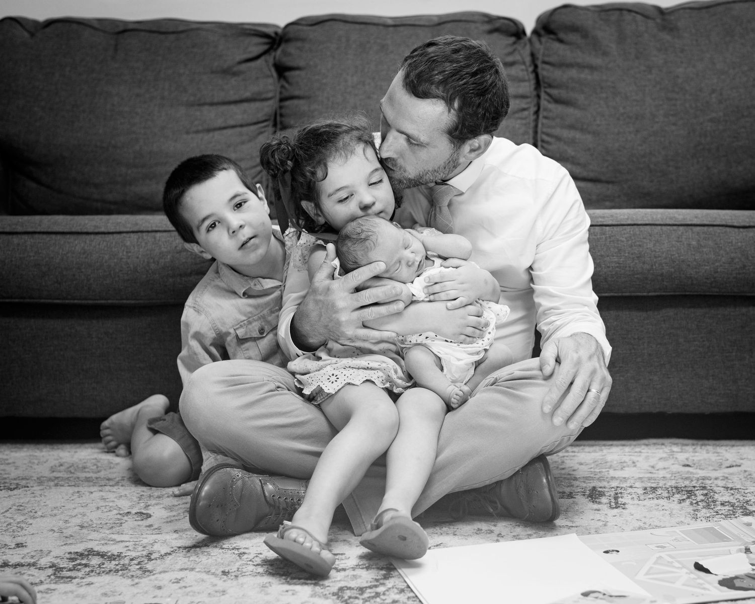 Emotional black and white photograph of big sister and big brother kissing a newborn with dad.