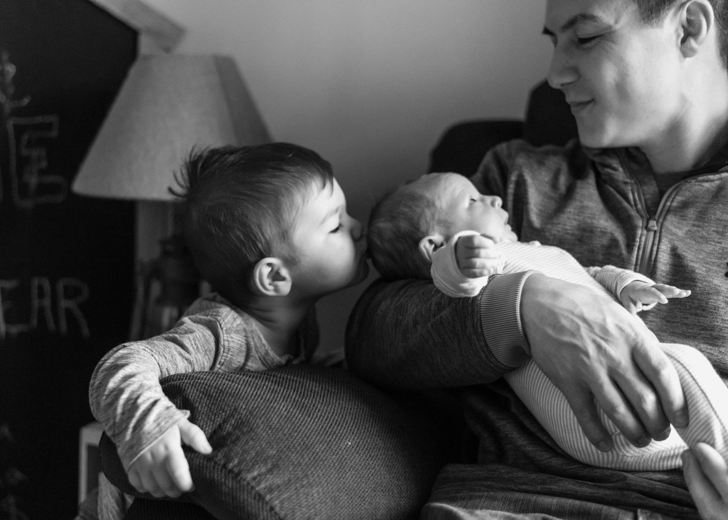 Toddler kisses newborn baby's head as dad watches.