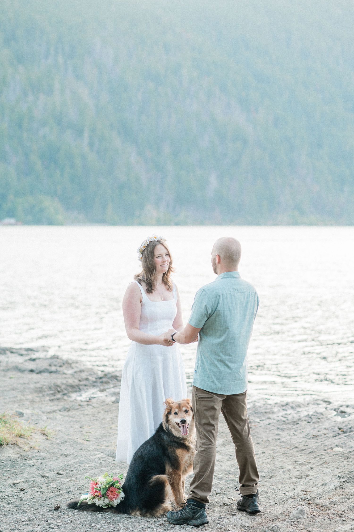 A couple shares a romantic moment on a lakeside beach with their dog sitting beside them during sunrise
