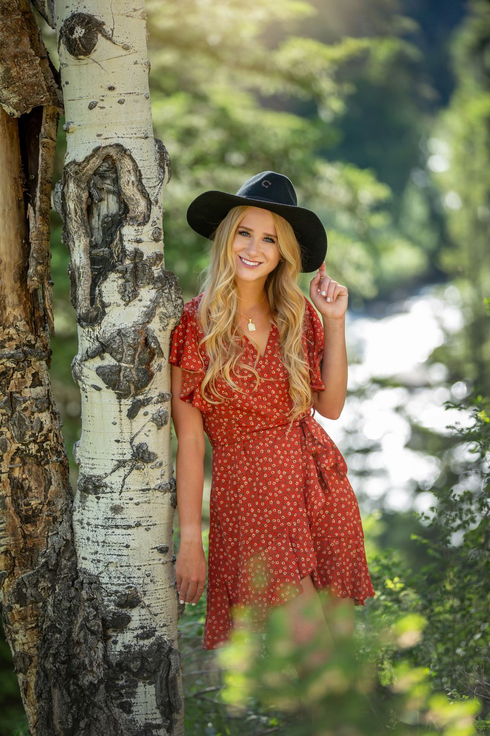 Smiling blonde woman in red floral dress and black hat leaning against birch tree in forest.