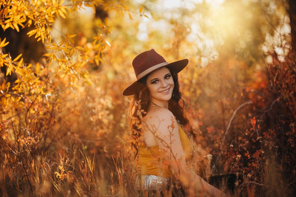 Young woman in brown hat smiles among golden autumn foliage in warm sunlight.
