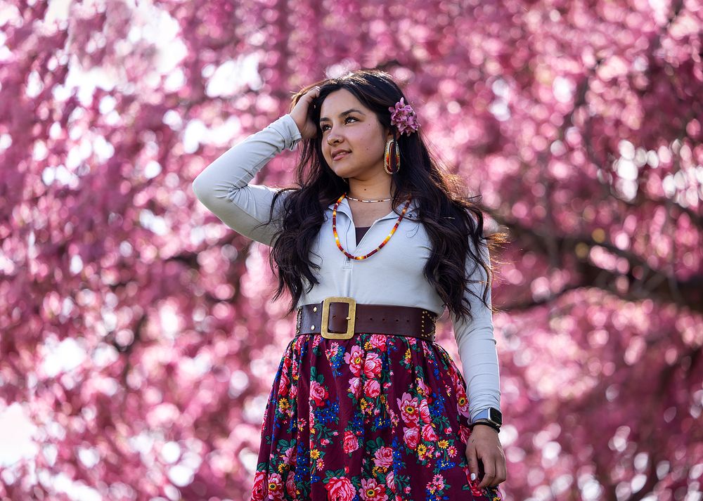 Young woman in a floral skirt poses gracefully beneath vibrant pink cherry blossom trees.