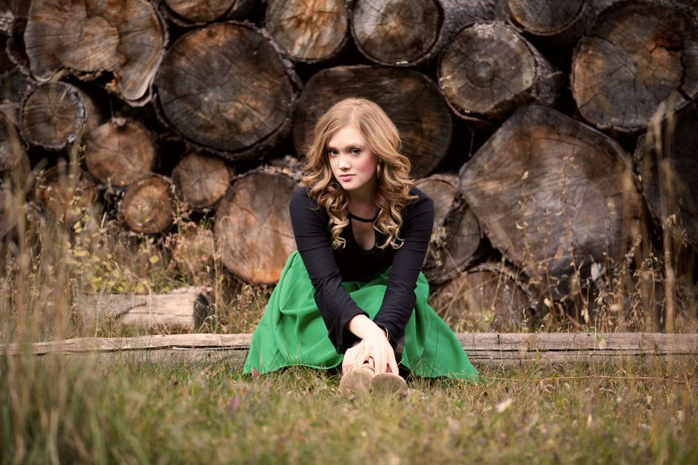Young blonde woman in black top and green skirt sitting outdoors near rustic log pile background.