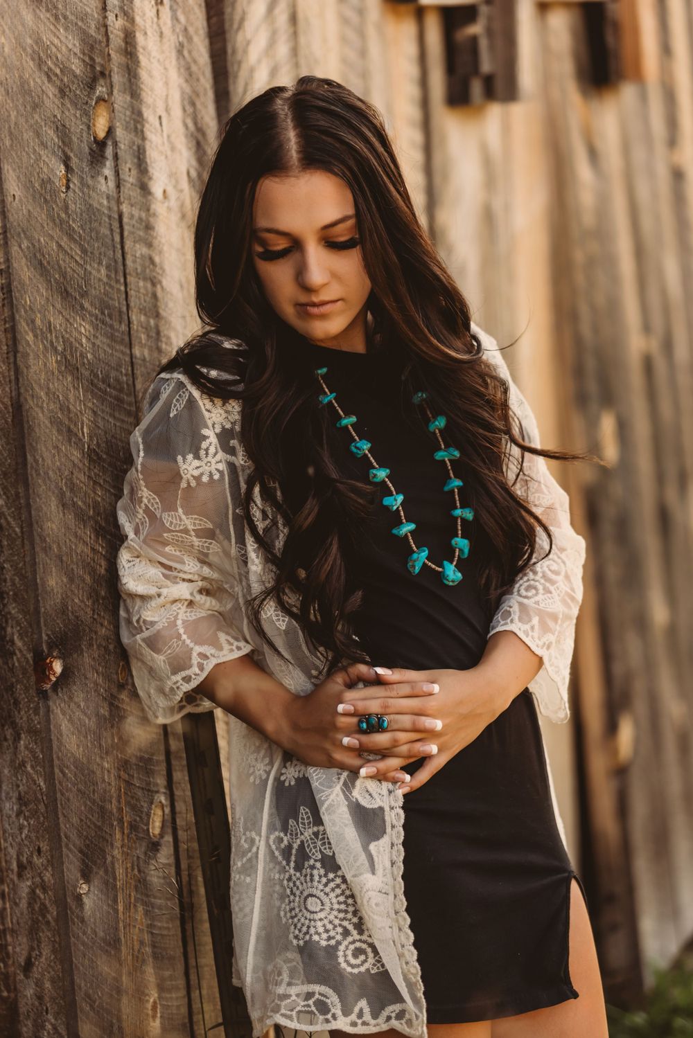 Brunette woman in black dress with turquoise necklace and lace kimono posing against weathered wood wall.