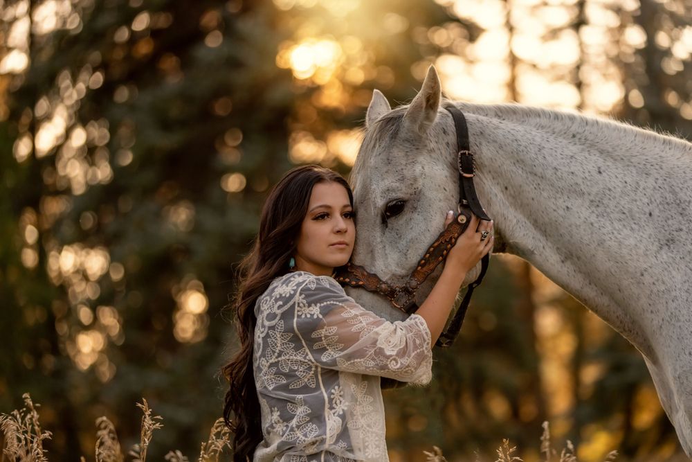 Young woman embracing a white horse at golden sunset in a forest setting.