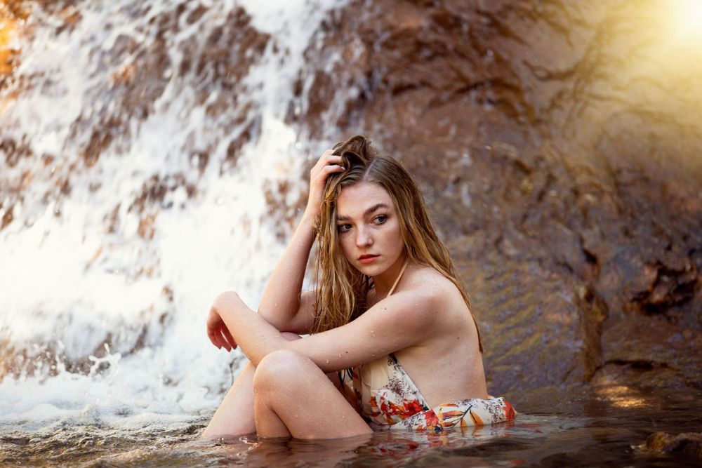 Young woman in floral swimsuit sits gracefully in shallow water before a sunlit waterfall.
