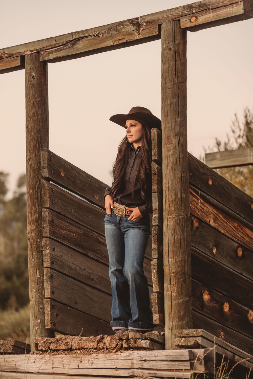 Cowgirl in brown hat and jeans standing on wooden ranch structure at sunset.