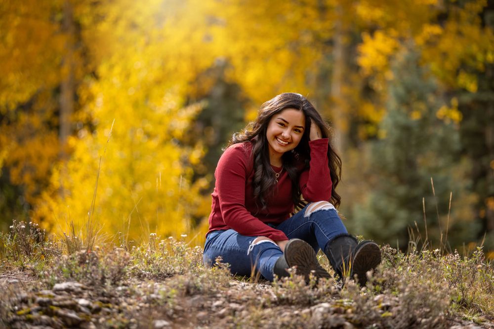 Smiling young woman in red sweater and jeans sitting outdoors among vibrant yellow autumn aspen trees.