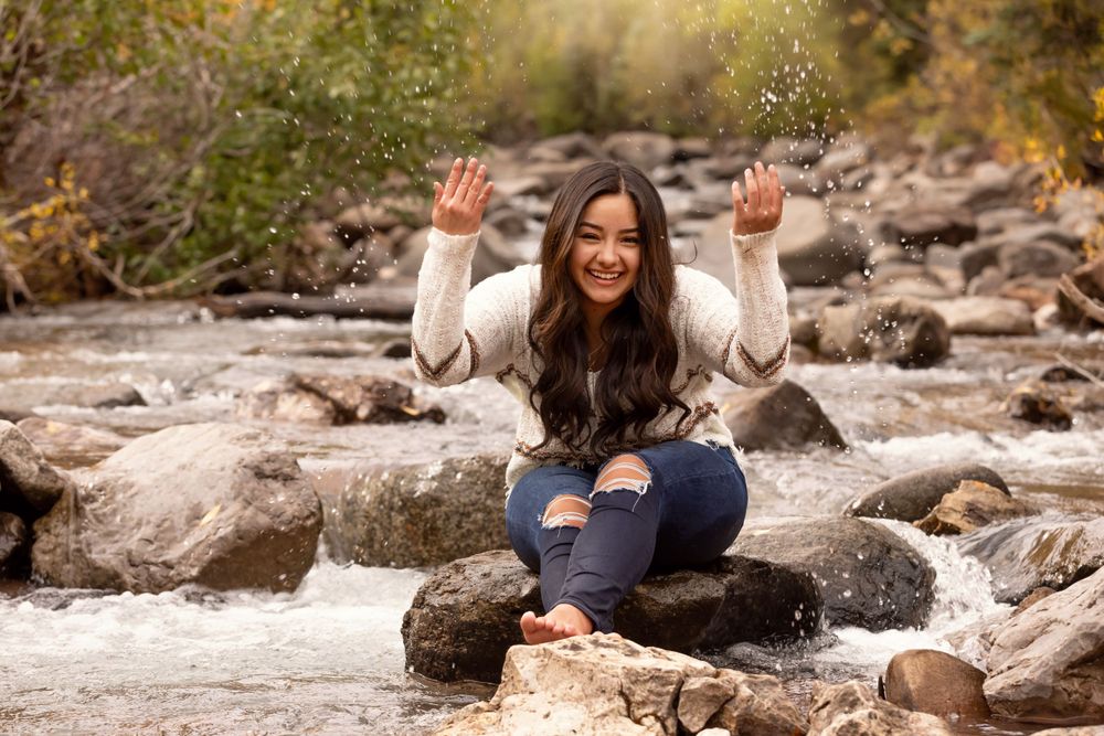 Young woman sitting on rocks by a stream, smiling and splashing water with both hands raised.