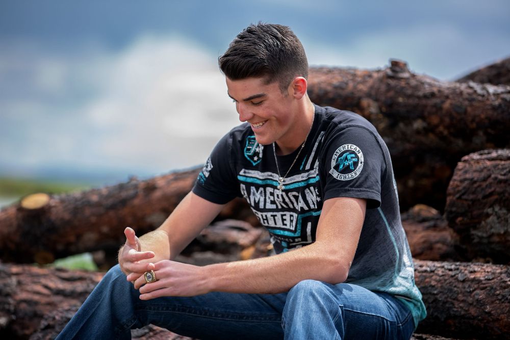 Young man in graphic tee sitting on logs smiling outdoors under cloudy sky.