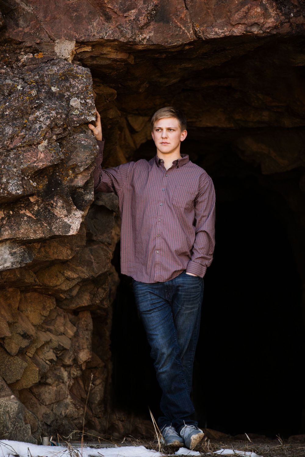 Young man in striped shirt leans against rocky cave entrance in a senior portrait photo.