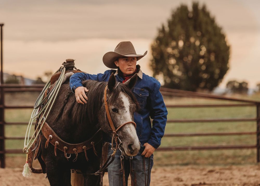 Young cowboy leading a dark horse in a ranch corral at dusk.