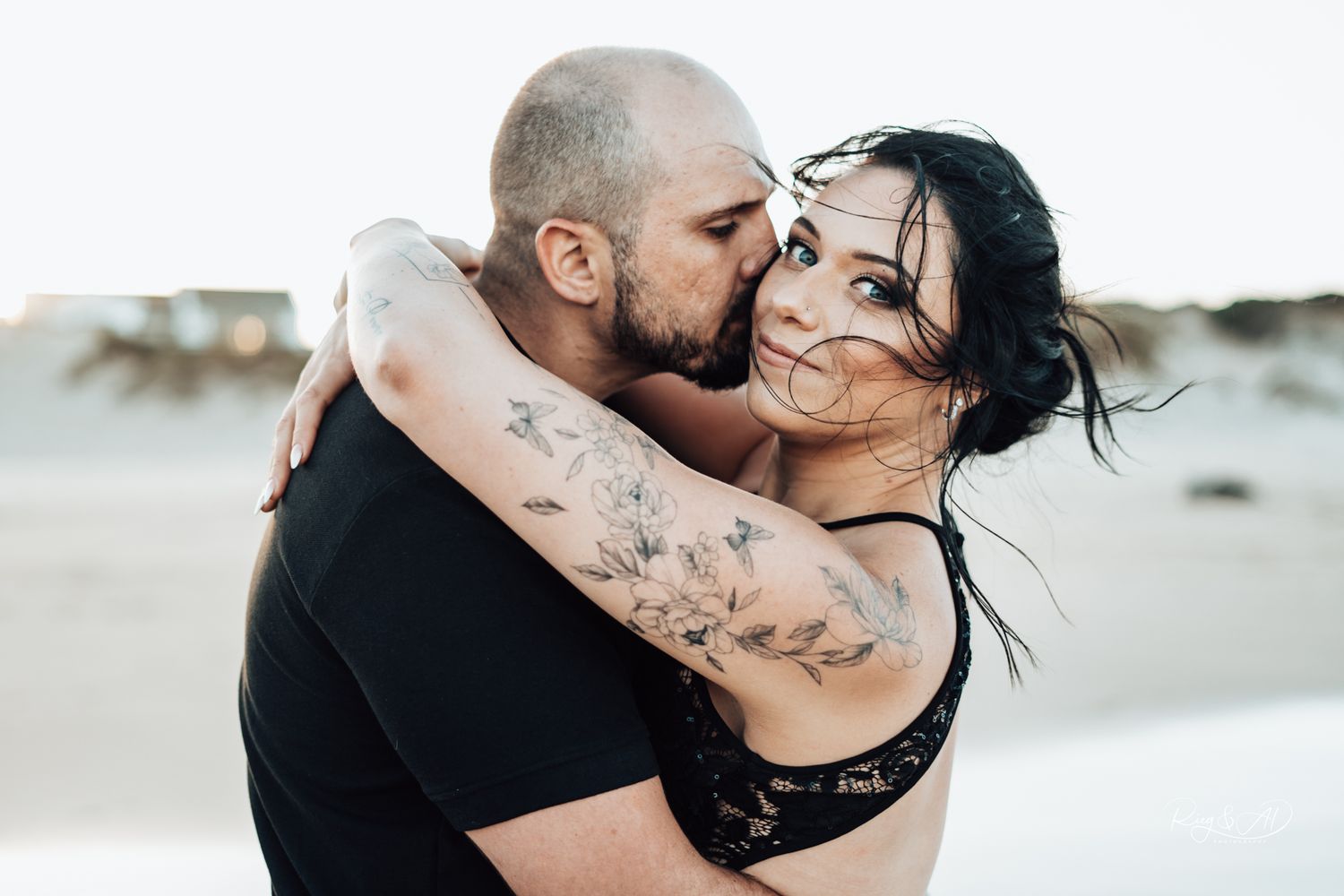 Tattooed couple shares an intimate moment on a sandy beach with the man kissing his partners cheek.