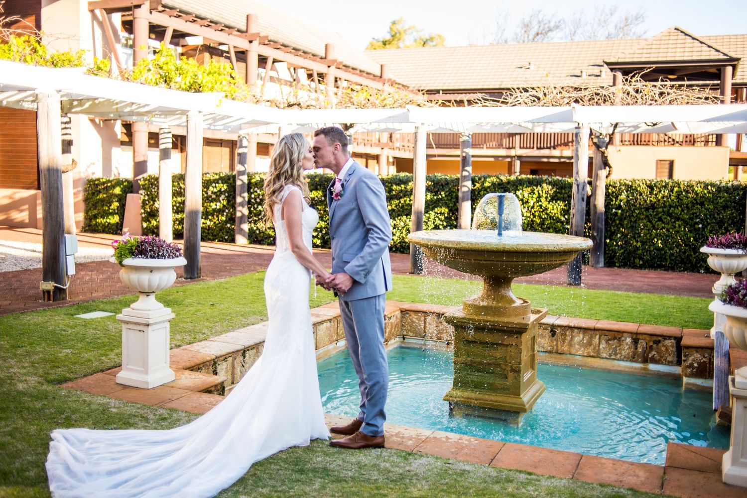 Couple poses by fountain in garden wedding venue with mountain backdrop.