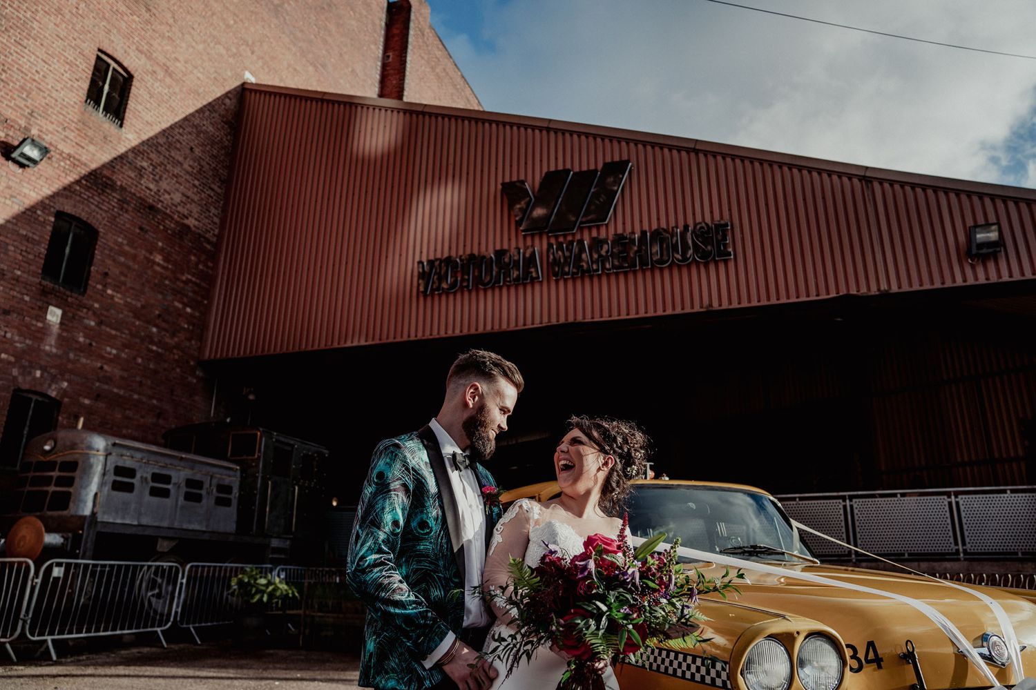 A stylish couple poses next to a vintage gold car outside a brick building.