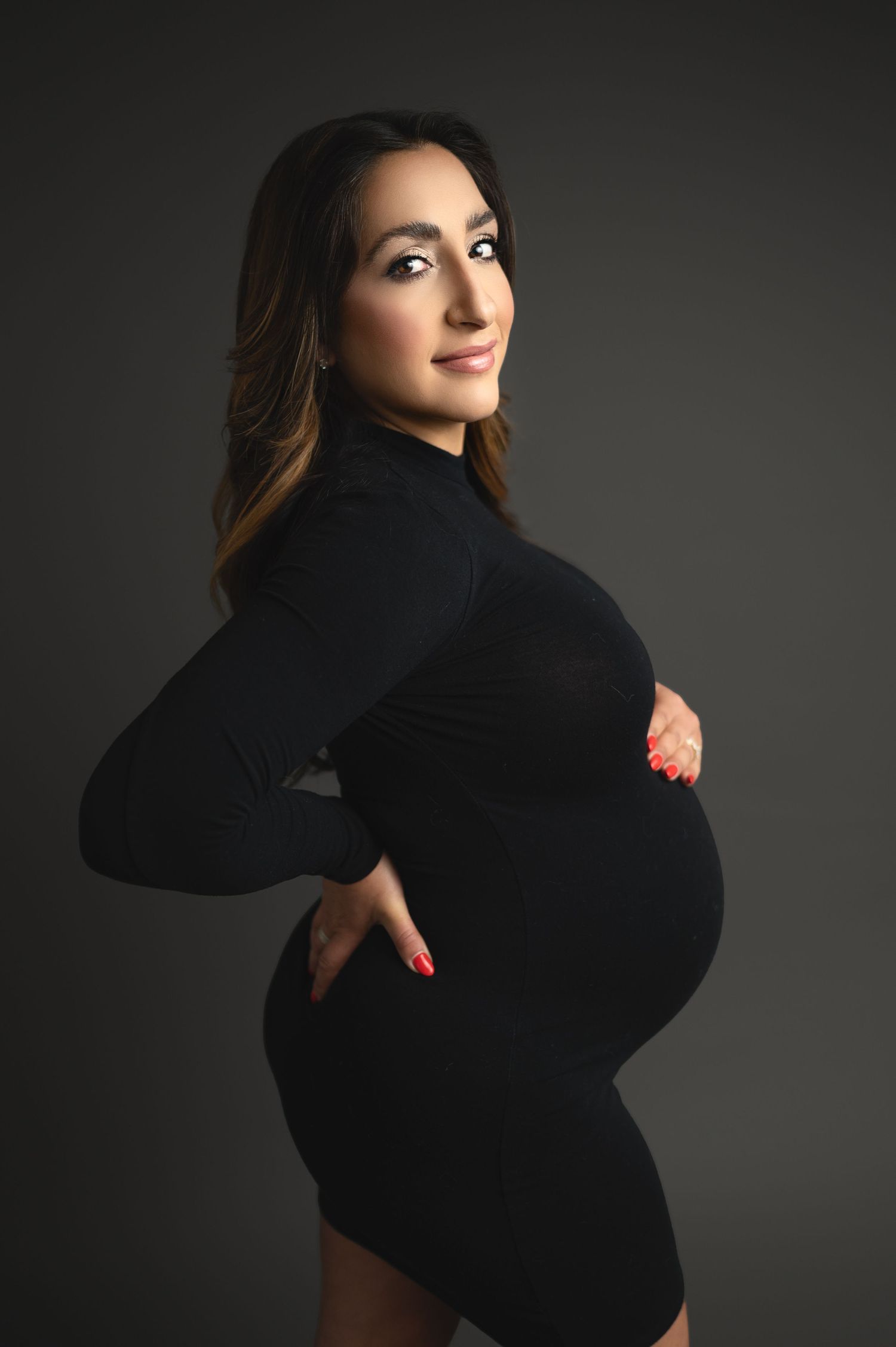 Elegant maternity photo of woman in black dress on dark backdrop.