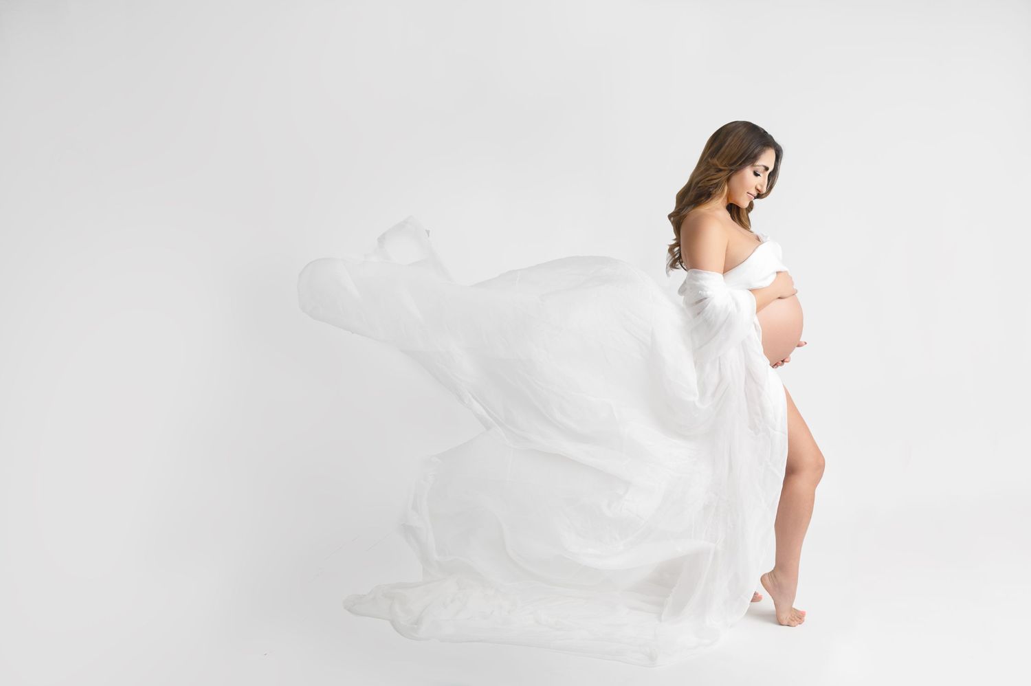 Ethereal maternity portrait with flowing white fabric in bright studio setting.