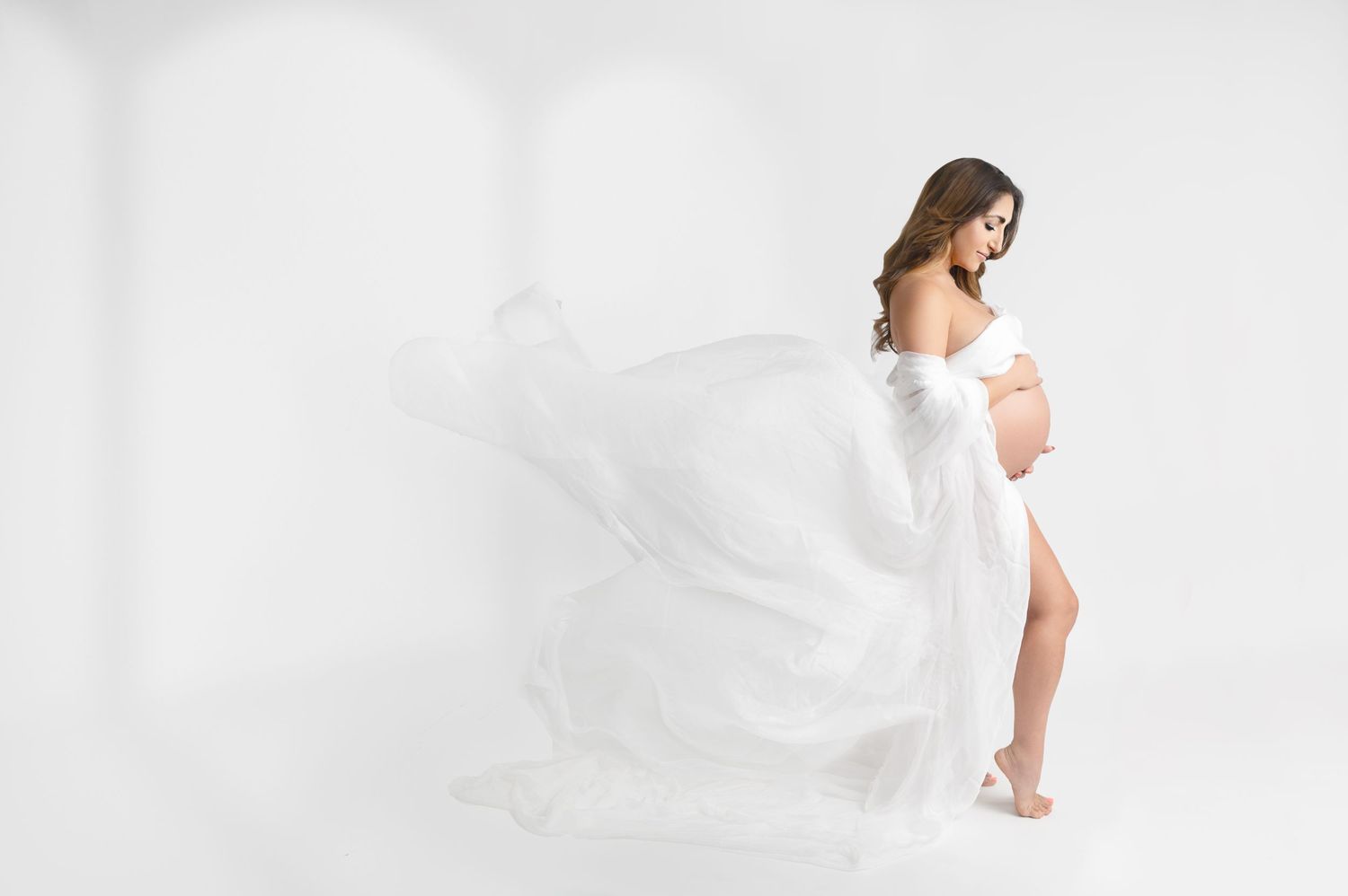 Flowing white fabric creates ethereal movement in elegant maternity portrait against white backdrop.