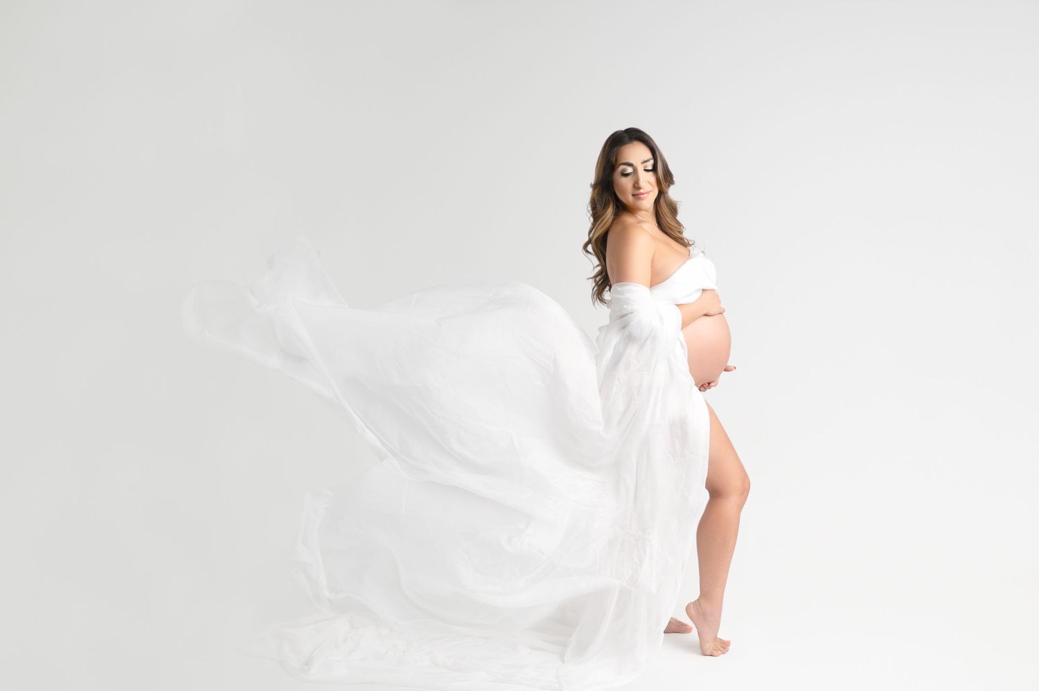 Ethereal maternity portrait features flowing white fabric creating dynamic movement in bright studio setting.