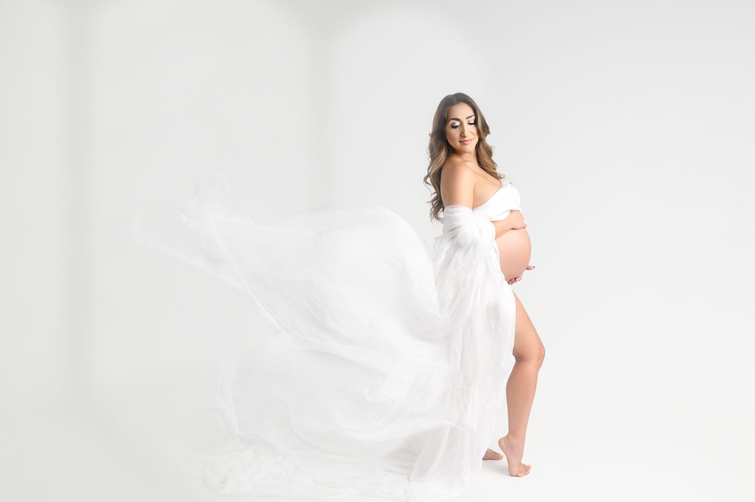 Maternity portrait with floating white fabric creates dreamy atmosphere against minimalist backdrop.