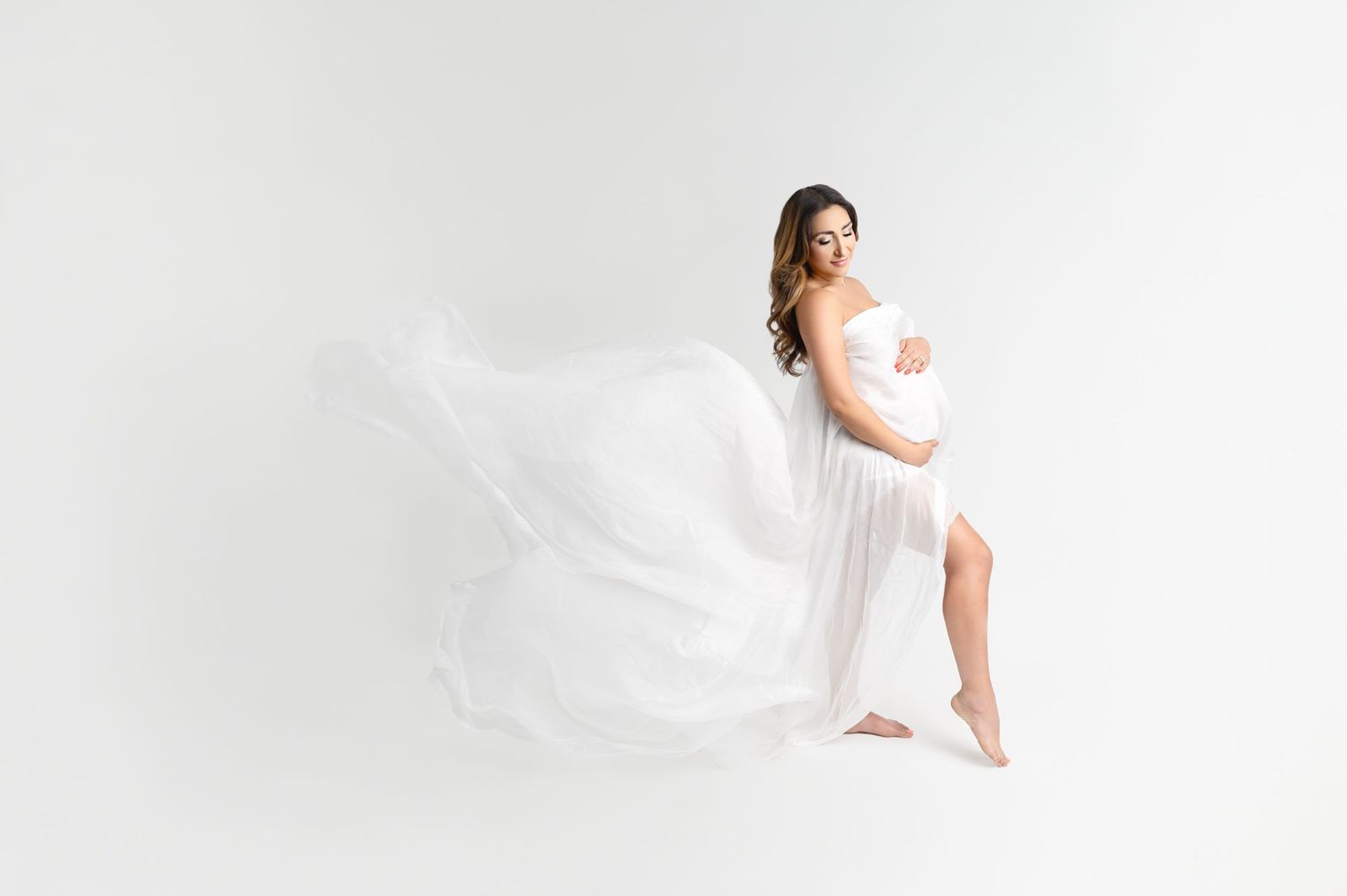 Elegant maternity portrait shows flowing fabric movement against minimalist white studio setting.