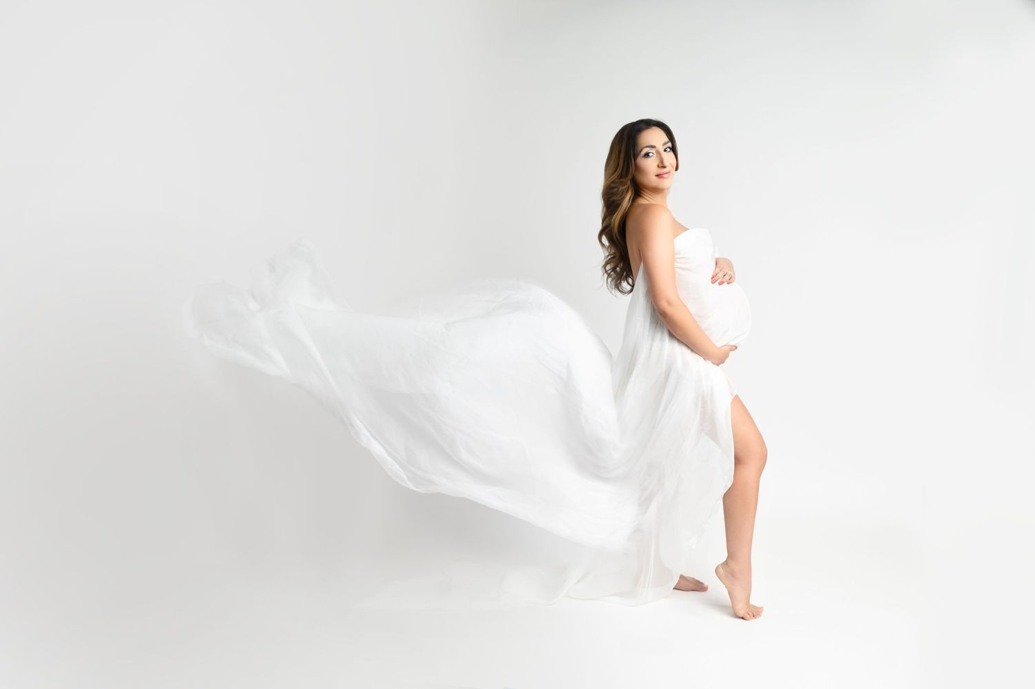 Woman in flowing white dress poses for ethereal maternity photo against white backdrop.