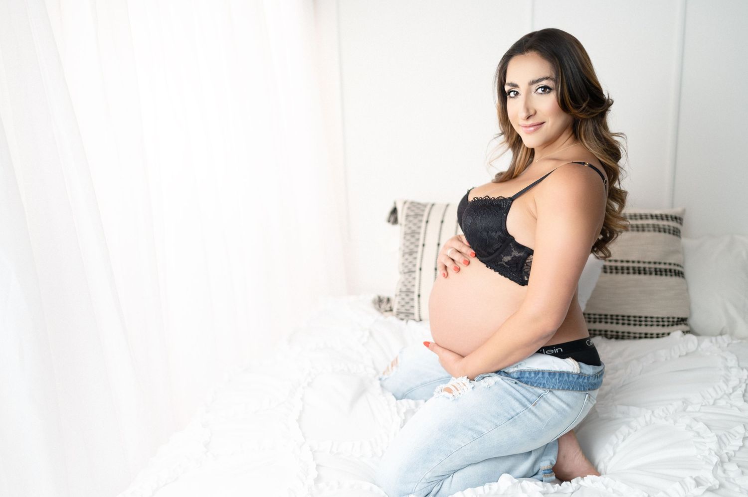 Pregnant woman in jeans and black bra poses on bed with decorative pillows.