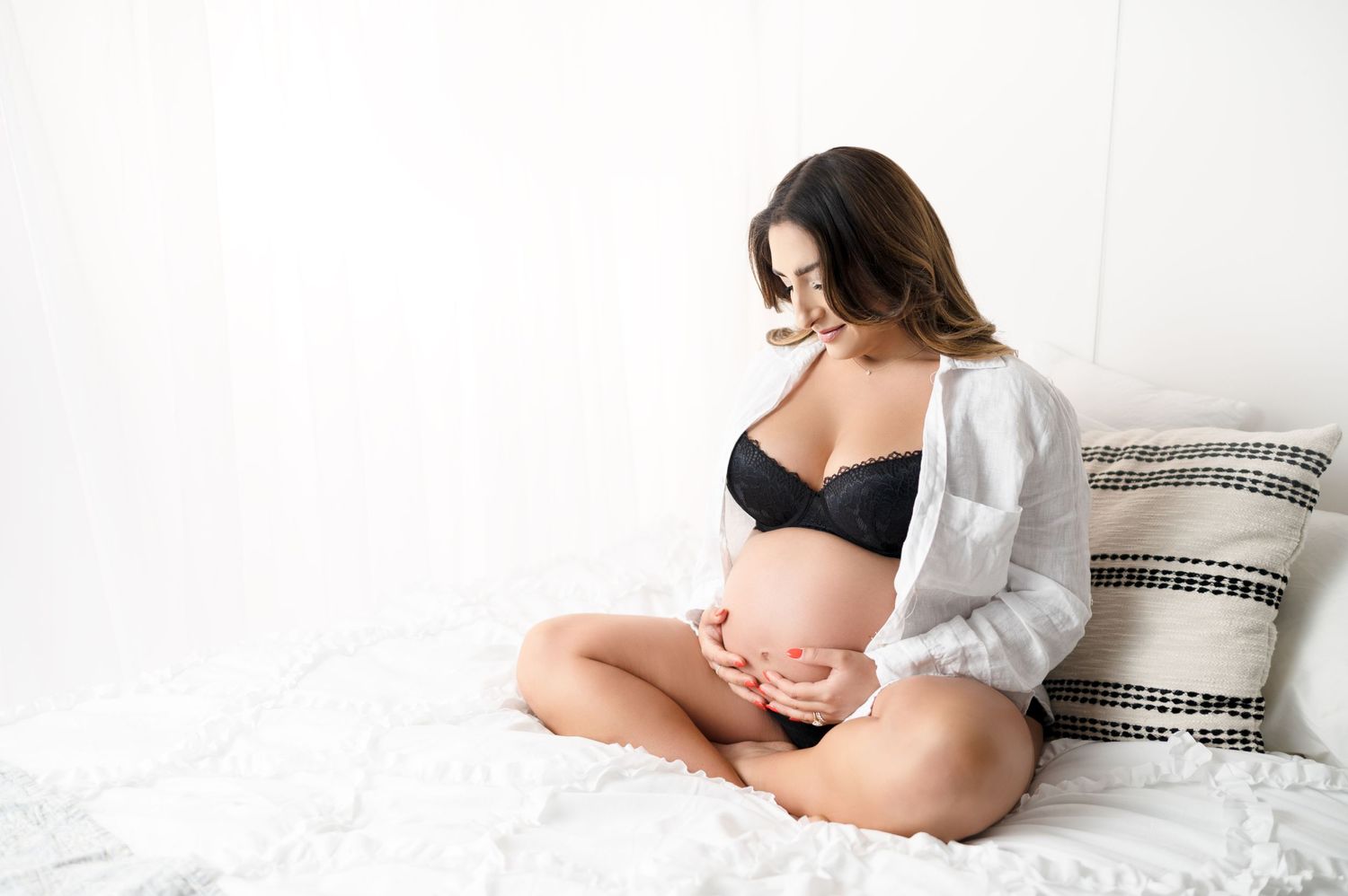 Mother-to-be in black bra and white cardigan sits on white bedding for maternity photos.