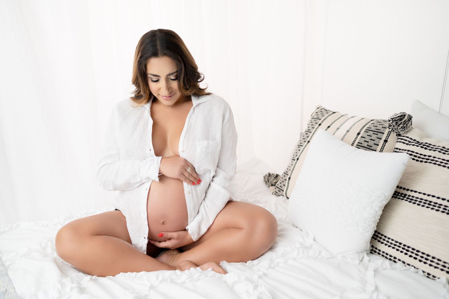 Expectant mother in white shirt poses cross-legged on white bed for maternity photoshoot.